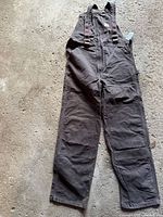 Full view of Carhartt dark brown workwear pants laid flat showing adjustable shoulder straps and front pockets.