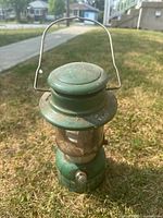 Green Coleman Easi-Lite camp lantern sitting on grass. Shows metal handle and rust spots on top and sides.