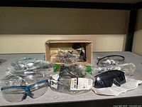 Photo showing assortment of safety glasses and sunglasses in plastic packaging and loose on shelf surface. Includes different frame colors: blue, black, clear, and darker tinted lenses