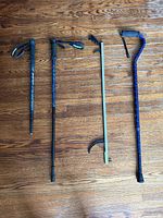 Four mobility aids laid flat on wooden floor: three walking canes with different handles and one reacher/grabber tool with claw and trigger handle.