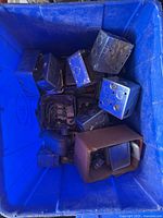 Top-down view of approximately a dozen metal electrical boxes in a blue container showing various sizes and internal wiring components in some boxes.
