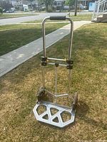 Full view of metal dolly/hand truck standing upright on grass, showing the foldable platform and handle with foam grip.