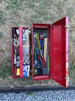 Open metal toolbox showing contents including various hand tools, ratchets, wrenches, screwdrivers, tape, and drill bits.