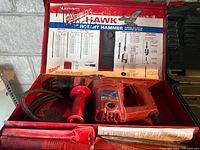 Milwaukee Hawk rotary hammer drill in red, inside open red metal case with drill specifications on lid, cord visible, with accessory packets.