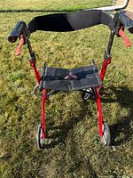 Front view of the red mobility walker showing the foldable black fabric seat, red frame, black handles with red handbrakes, and four wheels.