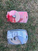 Front view of two Monster speaker cables in sealed clear plastic clamshell packaging on grass, one with red package label and one with blue package label.