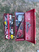 Red painted metal toolbox open showing assorted tools inside including hand saw, wrenches, pliers.