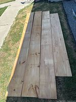Four long unfinished natural wooden boards laid out on grass and concrete.