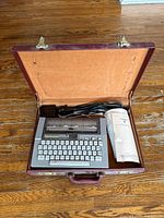 Open brown carrying case with Texas Instruments Silent 700 data terminal inside, alongside power cords and instruction manual.