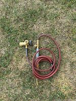 Full view of coiled red hose with attached metal torch handle and brass regulator on grass.