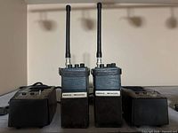 Two black handheld radios with long antennas standing upright between two dual-bay charging docks on a shelf, front view showing "MIKE WOOD" labels and control knobs on top of radios.