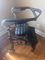 Side angle view of black mobility walker showing padded seat, ergonomic curved handles with hand brakes, black painted metal frame, and wheels.