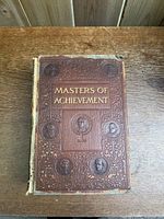 Front cover of the hardback book 'Masters of Achievement' showing embossed portraits and gold lettering.