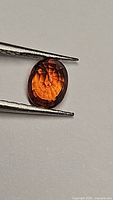 Close-up of oval-cut spessartite garnet held by tweezers on white background, showing bright orange color and internal clarity.