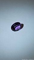 Close-up photo of deep purple oval cut amethyst stone on white background showing facets and color.