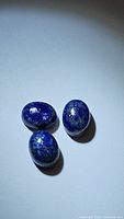 Three natural oval Lapis Lazuli stones arranged on a pale surface, showing deep blue hues and natural flecks.