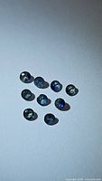 Ten round cut multicolored sapphires scattered on white background, showing varying blue and green hues and natural sparkle.