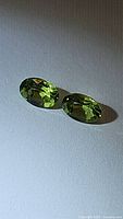 Pair of natural oval cut apple green peridot gemstones on white surface with shadow, shown from top view highlighting faceting and clarity.