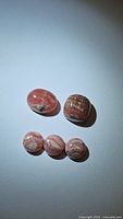 Five polished natural pink rhodochrosite stones arranged on a white surface with two larger stones at the top and three smaller round stones below.