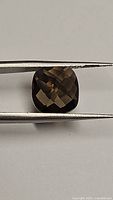 Top view of the natural smokey quartz gemstone showing the cushion shape and checkerboard facets under metal tweezers.