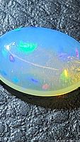 Close-up photo of oval cabochon opal showing vibrant flashes of blue, green, purple, and yellow colors on a milky translucent base.