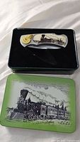 Folding cutter knife featured in black storage tray inside the decorative tin box with steam train design on the lid.