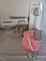 Three Pusheen themed bath items: stress ball in clear plastic packaging, bath brush with teal handle and Pusheen figure topper, pink bath pouf with Pusheen tag