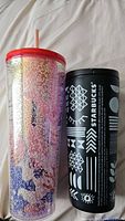 Pair of Starbucks tumblers, one tall clear glitter tumbler with colorful design and red lid with straw, one black tumbler with white geometric prints and Starbucks branding.