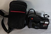 Black Pentax IQZoom 700 camera with lens cap and carrying case nearby, resting against a white wall