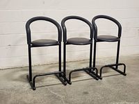 Front view of three black tubular metal counter-height stools with curved backrests