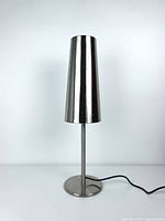 Front view of Talvik chrome silver table lamp showing tapered cylindrical metal shade and round base with power cable.