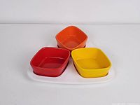 Set of three small square ceramic bowls in red, orange, and yellow, arranged on a white ceramic tray with three compartments, front view.