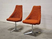 Front view of both chairs showing high backs, orange wool upholstery, and chrome bases against white cinderblock wall