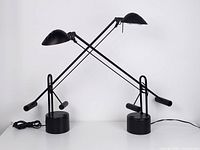 Front view of two black vintage Stilplast table lamps with counterweight arms crossed on a white background.