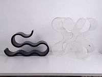 Both wine bottle holders side by side on white surface against white background. One transparent with curved six-section design, the other black with wavy shape.