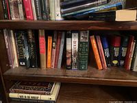 Two shelves of a wooden bookshelf filled with various hardcover and paperback books, upright and some horizontally stacked.