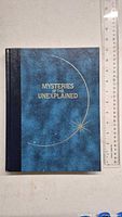 Front view of the Mysteries of the Unexplained hardcover book showing blue textured cover with crescent and star design and gold embossed title.
