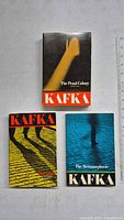 Top view of three Kafka paperback books arranged on a grey surface: The Penal Colony at top center, The Trial at bottom left, and The Metamorphosis at bottom right.