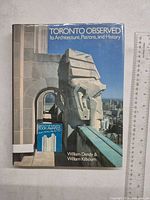Front view of the book with dust jacket showing architectural sculpture close-up and Toronto skyline background, with partial ruler for scale.