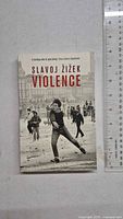 Front cover of 'Violence' by Slavoj Žižek showing a black and white photo of a street protester tossing an object, with a ruler for dimension reference.