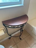 Photo showing half moon accent table with dark wood top and black metal scrollwork frame placed against wall below window.