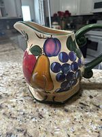 Front view showcasing fruit designs on the ceramic water pitcher including grapes, pear, and apple with green handle.