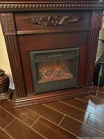 Front view of dark wood electric fireplace with detailed carvings and electric firebox showing ember effect on a wooden floor.