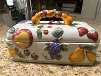 Front view of the ceramic bread basket lid showing colorful fruit decorations including peaches, cherries, strawberries, grapes, and pears with a handle designed to look like fruit clusters.