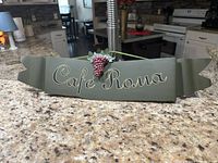 Front angle of metal wall hanging on granite countertop showing 'Cafe Roma' text and grape cluster