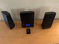 Front view of mini stereo system showing main unit with digital display, two separate speakers, and remote control on surface.