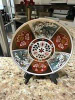 Front view of an oriental china display plate featuring floral and phoenix patterns in red, gold, green, and blue colors on a beige fragment patterned background.