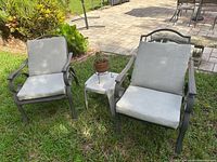 Front view of two metal patio chairs with grey cushions and small white side table with potted plant on a paved patio.