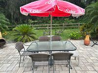 Rectangular glass-top patio table with six woven chairs and Coca-Cola branded red umbrella open on top in outdoor patio area.