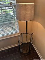 Floor lamp with beige fabric shade positioned on a round glass side table with black metal frame near window and wall corner.